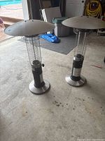 Two tabletop propane patio heaters with cylindrical bodies, metal wire cages, and round metal reflector tops standing on a concrete floor in a garage setting.