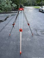Full view of the aluminum survey tripod standing on asphalt, showing telescoping legs with orange feet and clamps for height adjustment.