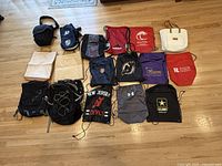 All 16 bags arranged on floor including Eddie Bauer backpack, camera bags, beach bags, and drawstring gym sacks with various colors and logos