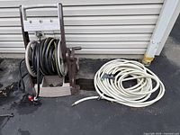 Wide shot showing the garden hose reel with black hose and coiled white hose beside it, outdoors on pavement near siding.