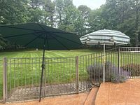 Two patio umbrellas set up outdoors near a fence; one solid green and one green and white striped with decorative pattern.