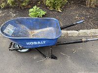 Side view of the blue Kobalt metal wheelbarrow with single pneumatic tire on pavement, showing dirt and wear.