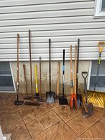 Photo showing 13 assorted yard and garden tools leaning against a wall on a paved surface, including hoes, rakes, shovels, post hole diggers, an axe, and a snow shovel, in used condition with some surface rust and wear.