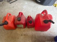 Four red plastic gas cans of varying sizes arranged side-by-side on concrete floor, showing overall wear and dirt