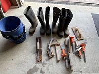 Full lot overview showing three pairs of black rubber boots, blue bucket, and various masonry hand tools arranged on a concrete floor.