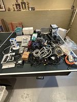 Wide view of mixed electronics and accessories spread over a ping pong table showing various cables, power strips, devices, and packaging.