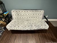 Full view of futon sofa bed with geometric-patterned gray fabric mattress cover and black metal frame in room with hardwood floor.