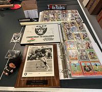 Photo showing a signed Tim Brown Oakland Raiders plaque and several pages of football cards in a binder with additional Raiders memorabilia items on display