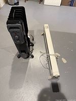Photo showing black oil-filled radiator heater and white electric baseboard heater side by side on floor, cord visible on baseboard heater.