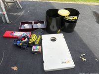 Full overview of the collection of outdoor games including cornhole board, Kan Jam bins, Spikeball bag, bocce balls case, horseshoe frames, and chalk box on pavement.