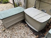 Two outdoor plastic deck boxes side by side on gravel under outdoor setup showing general condition and color contrast.