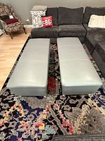 Two rectangular light gray leather or faux leather ottomans placed side by side on a multi-colored patterned rug in front of a dark sectional sofa. Ottomans show smooth surfaces and some stitching detail.