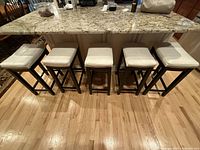 Photo showing five counter stools with white faux leather padded seats and dark wooden legs lined up beneath a granite countertop.