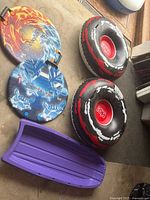 Five snow sleds and tubes arranged on a carpet, including two circular Snowbuggy sleds with fire and blue ice designs, two black and red inflatable tubes branded K2 with white accents, and a purple molded plastic sled.