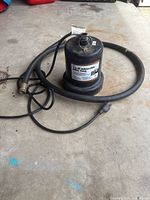 Full view of the black cylindrical electric submersible pump sitting on concrete floor with black attached power cable and hose coiled around it.
