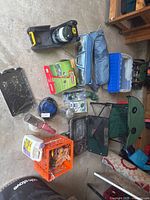 Camping gear items arranged on garage floor including tents, Coleman lantern, stoves, grill, fog machine, dinnerware, griddle, coffee pot, shovel, fishing rod, collapsible table, and more