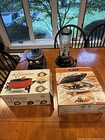 Photo of all five items on wooden table including boxed West Bend Fondue Pot and Bella Pie Maker, Cooks Waffle Maker, Osterizer Blender, and BonJour Torch