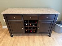 Front view showing three drawers on top, two cabinet doors on sides, and central wine rack with multiple wine bottles inside. Marble top is flat and glossy with light color tones.