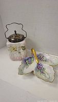 View of antique ceramic canister with floral and bird decoration, metal lid and wire handle. Also shows three-section leaf-shaped Limoges dish with purple floral decoration and gold handle.