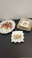 Set of five assorted porcelain plates and dishes on black surface, showing fruit, floral, and royal commemorative designs