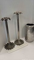 Two tall silver plated candle holders with decorative leaf-shaped bases and tops, placed side by side with a large silver-colored hammered texture vase behind, and two white glass candle holders lying in front.