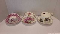 Front view of three tea cup and saucer sets, showing floral patterns and crown emblem designs, arranged side by side
