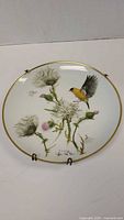 Front view of round collector plate with painted goldfinch bird among thistles and gold rim