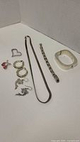 Photo showing three pairs of earrings (pink teardrop, silver hoop, silver bird), silver-tone chain necklace, silver link bracelet, white bangle, and heart-shaped pendant.