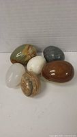 Photo of all six polished stone eggs arranged on a flat surface showing varied colors and patterns including green, gray, white, and brown tones.