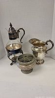 Silver plate and glass carafe on stand, silver plate pitcher, and small vase shown together on white background.