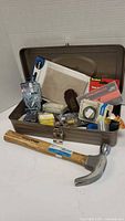 Metal toolbox open showing a collection of small tools and supplies including a hammer, tape measure, nails, screws, and tape rolls.
