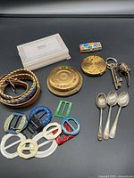 Overview of lot displaying silver spoons, vintage compacts, scarf belt buckles, beaded belt, trinket box, keys, and pin