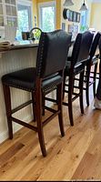 Three of the four bar stools aligned along a kitchen island, showing front and side views of dark brown faux leather upholstery, nailhead trim, and wooden frames.