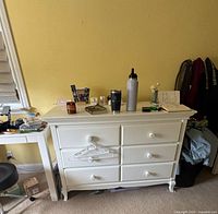 White dresser with six drawers containing various personal items on top such as cups, water bottle, and papers. Room has yellow painted walls.