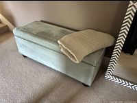 Blue fabric covered rectangular trunk with copper studs along the edges, shown closed and resting on carpet beside a mirror and wall.
