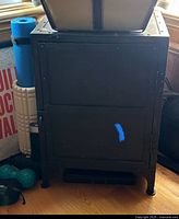 Closed black metal cabinet with visible riveted edges and short legs, surrounded by various household items.