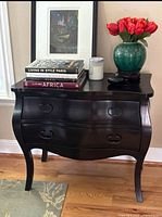 Full view of brown 2 drawer wooden chest with decorative metal handles, curved legs, placed on wooden floor with books, vase, candle on top.