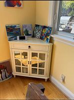 White wooden corner cabinet positioned in a room corner with three colorful paintings on top and a digital clock.
