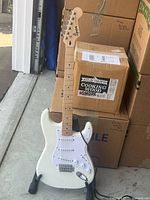 White Squier Stratocaster guitar on a black folding stand with cardboard boxes background