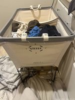 Side angle of the Steele French Laundry Basket filled partially with clothes showing the Steele branding and metal frame.