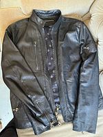 Full view of black leather jacket with front zipper, zipped chest pocket and zipped sleeve pocket.