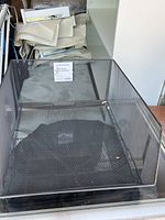 Front view of a large graphite metal mesh stacking basket with square base and tall mesh sides, placed on a glass surface. Basket contains a label.
