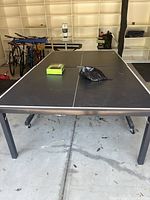 Full view of the black ping pong table with white lines on the surface placed in a garage with bicycles in the background.