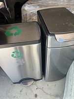 Two Simple Human stainless steel bins positioned side by side, one with recycling logos and one trash can with foot pedal, visible wear and use marks.