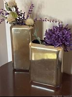 Two gold metal vases with an assortment of artificial flowers, shown from the front on a wooden surface.