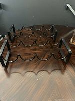 Two black metal stacking wine racks on a wooden surface, each with 6 curved slots to hold wine bottles horizontally.