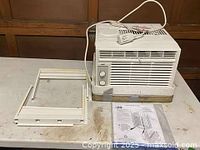 Photo showing the white window air conditioning unit placed on a surface with attached power cord, the white mounting frame and instruction manual placed next to it.