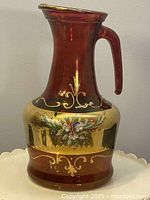 Front view of the cranberry glass pitcher showing gold enamel scrollwork and hand-painted floral design.