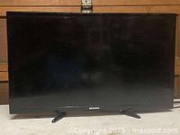 Front view of black Sylvania flat screen television with dual legs, screen off showing damage or broken display.
