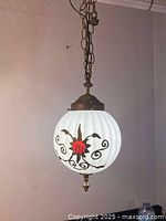 Full view of the hanging lamp installed on the ceiling, showing the white ribbed glass globe with dark metal floral and scroll embellishments and red flower accent
